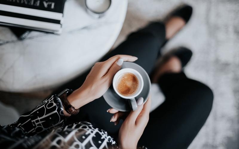 business woman sits next to a marble table with books and holds a saucer with a cup of espresso